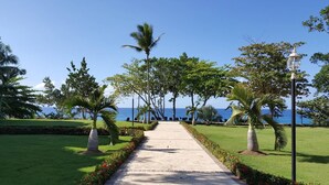 A paved pathway lined with palm trees and shrubs, leading to a view of the ocean. - Premium 3-BR Ocean view Condominium at Hispaniola Beach! (Sosúa)