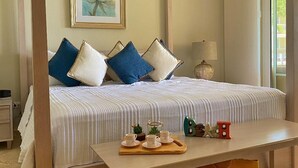 A bedroom with a large bed, a bedside table with a tray of tea cups, a framed picture on the wall, and a window with a view of greenery. - Premium 3-BR Ocean view Condominium at Hispaniola Beach! (Sosúa)
