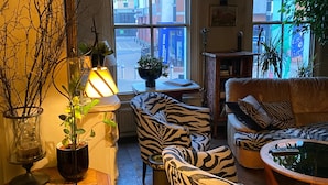 Ein gemütliches Wohnzimmer mit Sesseln in ZEBRA-Muster, einem Couchtisch und einer Lampe. Fenster bieten Blick auf Gebäude.