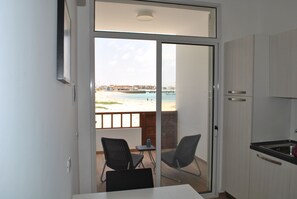 Varanda com vista para a praia e mar, uma mesa, duas cadeiras e uma mesinha com um objeto azul.