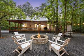 A cabin with a stone fire pit surrounded by white chairs, set in a forested area with a deck and outdoor seating.