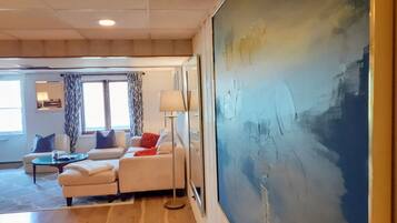 Un salon avec un grand tableau abstrait, un ensemble de meubles de salon, une lampe et une vue sur une pièce avec une fenêtre et des rideaux.