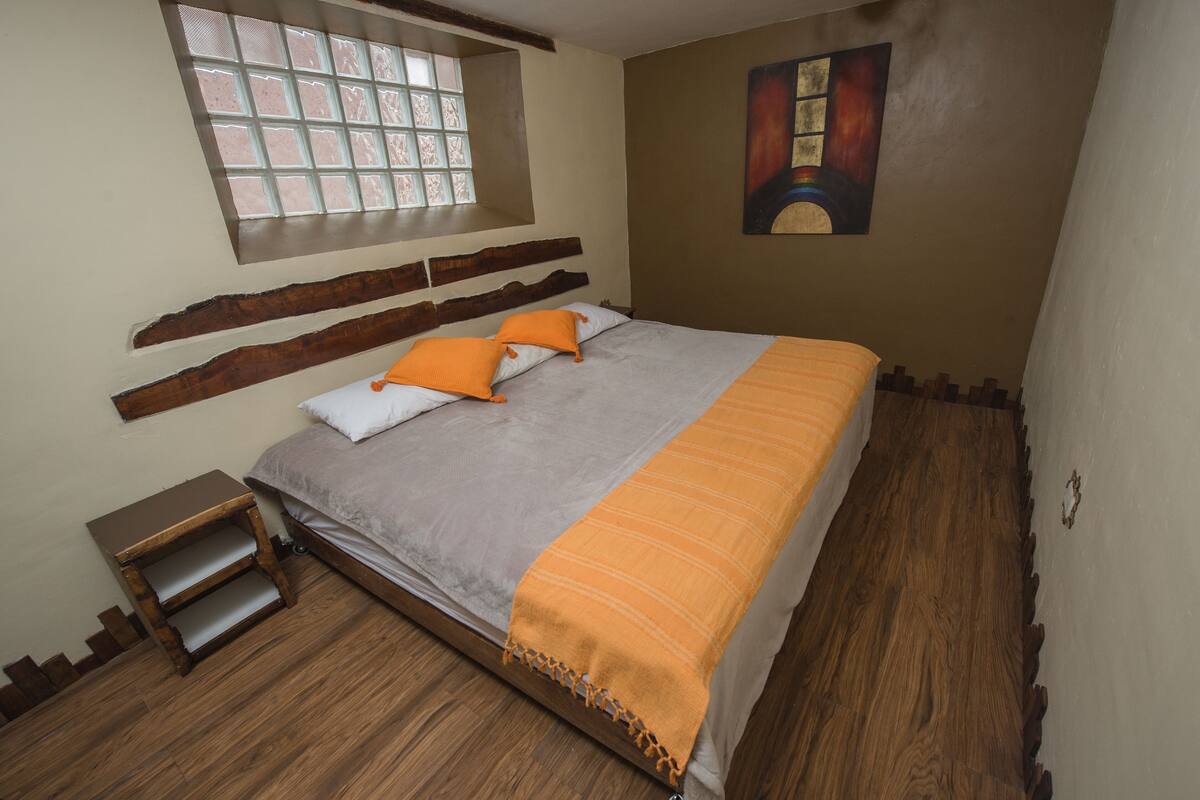 Un dormitorio con cama, cabecero de madera, una ventana, un cuadro y un taburete de madera.