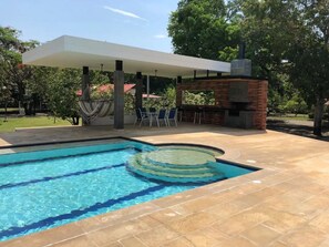 Una piscina con una zona circular hundida, un área de descanso cubierta y una estructura de ladrillo con chimenea.