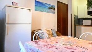 A dining area with a table covered in a bakery-themed tablecloth, a refrigerator, a painting of a beach, and a kitchen area with cabinets and a microwave. - N040 - Numana, three-room apartment a stone's throw from the center (Numana)