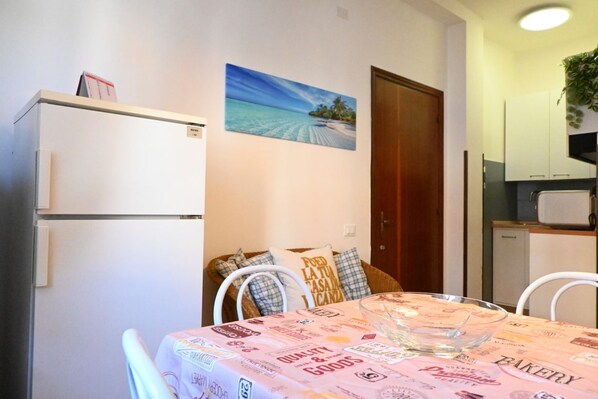 A dining area with a table covered in a bakery-themed tablecloth, a refrigerator, a painting of a beach, and a kitchen area with cabinets and a microwave.