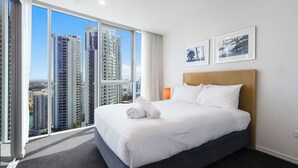 A hotel room with a large bed, white bedding, and a view of tall buildings through floor-to-ceiling windows.