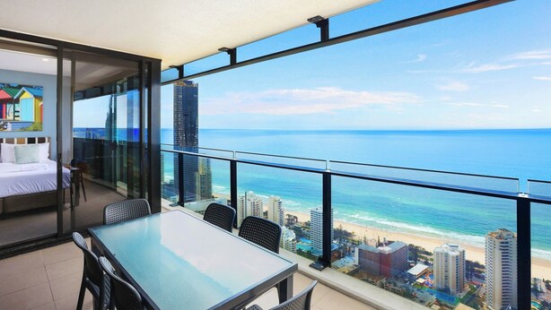 A balcony with a glass door leading to a bedroom with a bed and a table. The balcony overlooks a beach and cityscape.