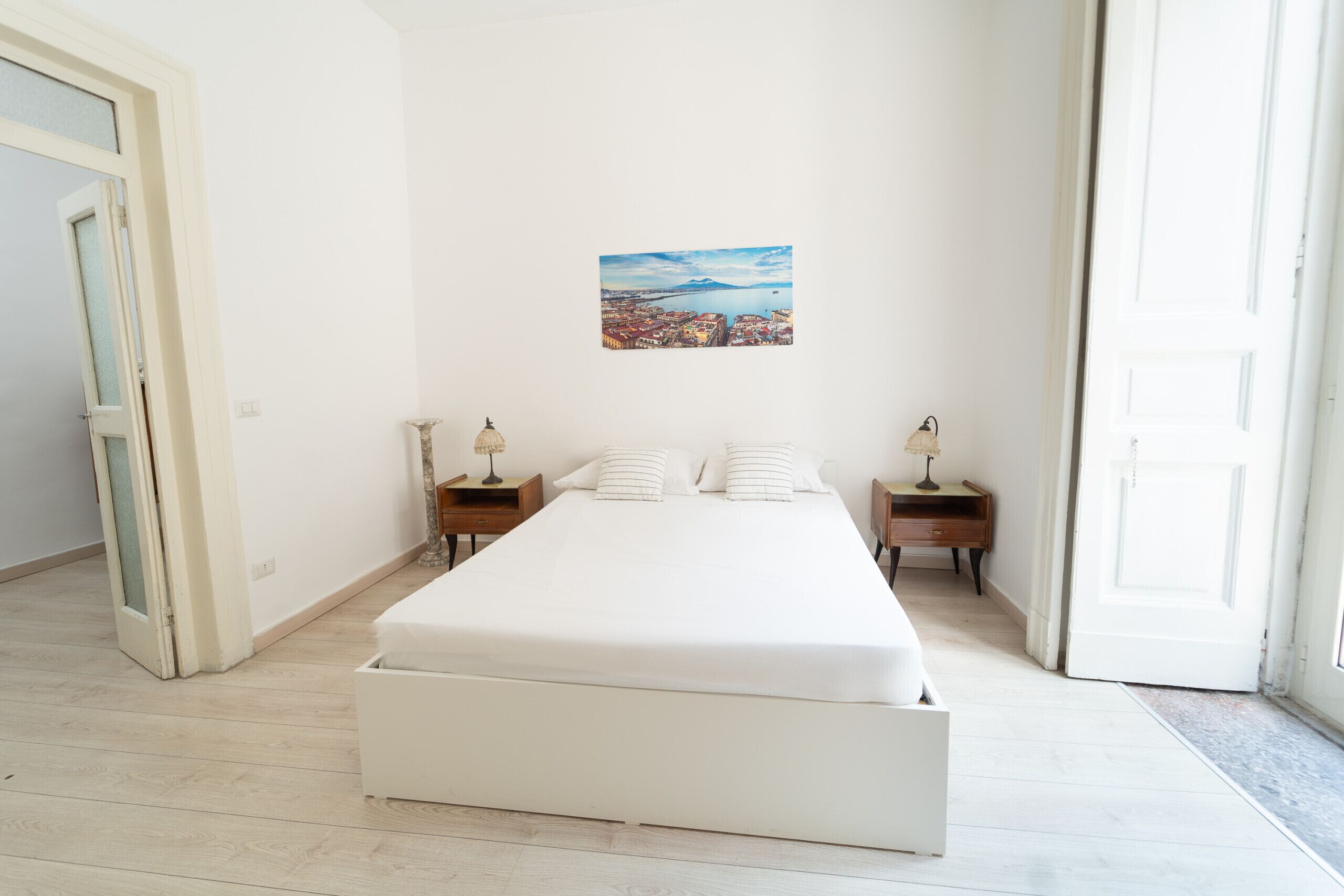 Ein Schlafzimmer mit einem großen Bett, zwei Nachttischen, einem Wandbild und einer Tür zu einem anderen Raum.
