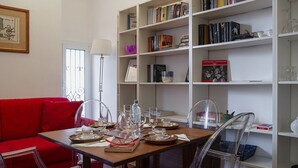 A dining area with a wooden table set for four, surrounded by clear chairs. A red sofa is against the wall, and a white bookshelf filled with books and decorative items is to the right. - Cozy Family Apt in Castelletto by Wonderful Italy (Genova)