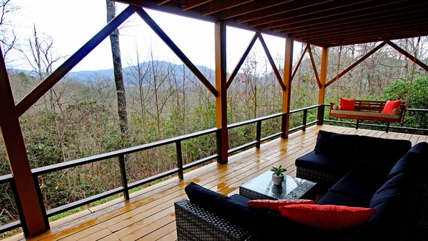 A covered outdoor seating area with wicker furniture and a bench with red cushions, overlooking a forested landscape. - Mountain Views & Solitude. This is Shadow Ridge. Hot Tub. (Fletcher)