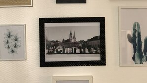 Een muur met diverse kunstwerken in lijsten, waaronder bloemenmotieven, een stadsgezicht en een cactuss.