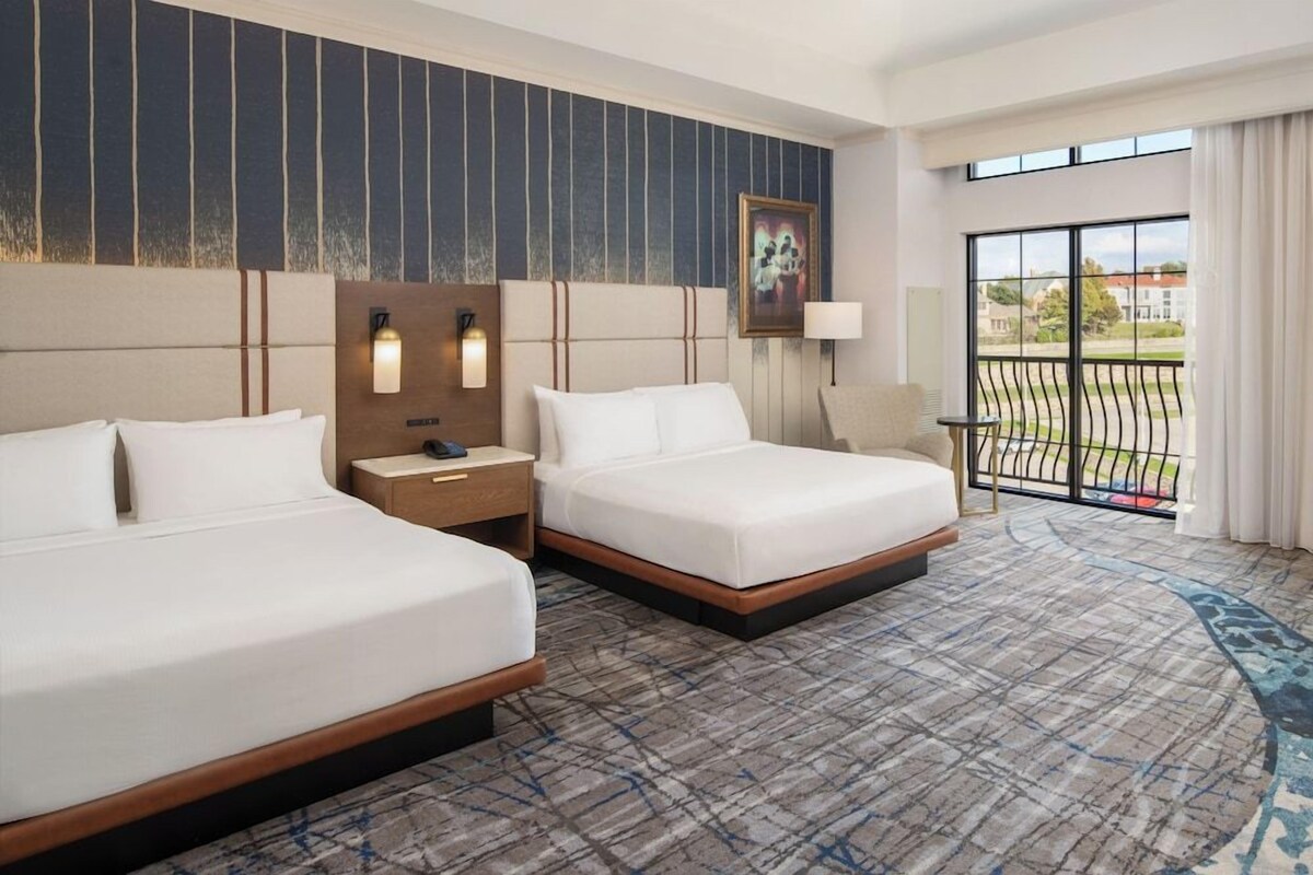 A hotel room with two beds, a nightstand, a lamp, a painting, and a balcony with a view.