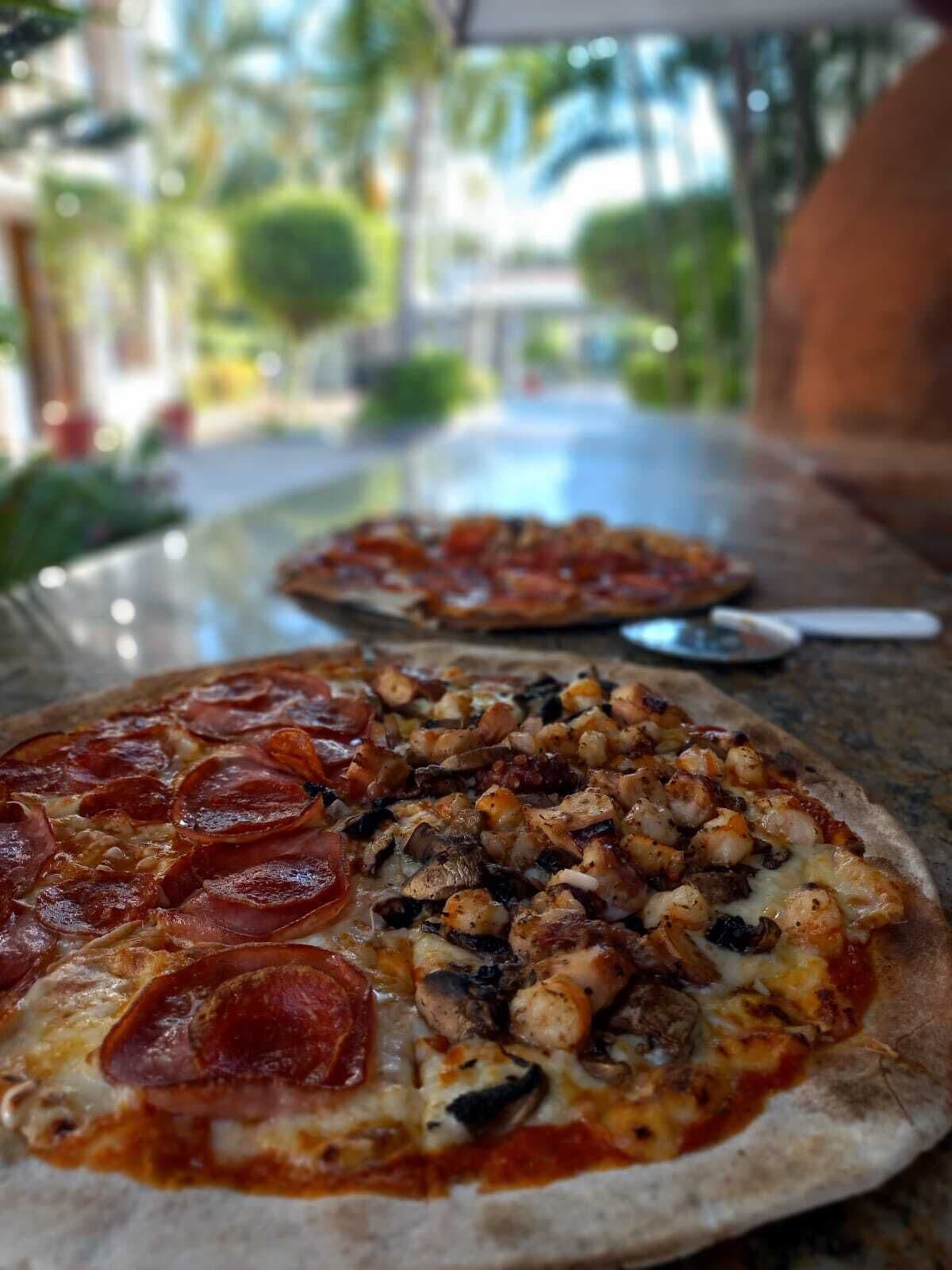 Two pizzas on a table with a view of greenery and a building in the background.