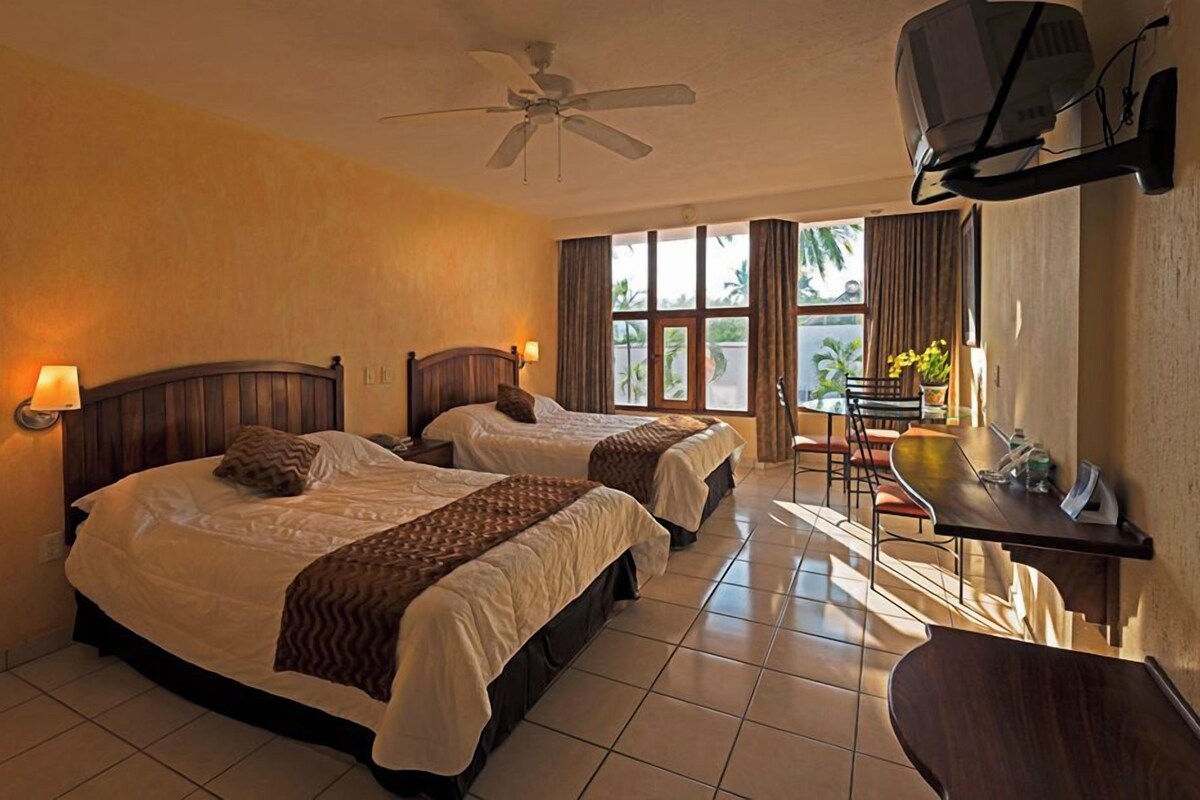 A hotel room with two beds, a desk, a chair, a television, and a ceiling fan.