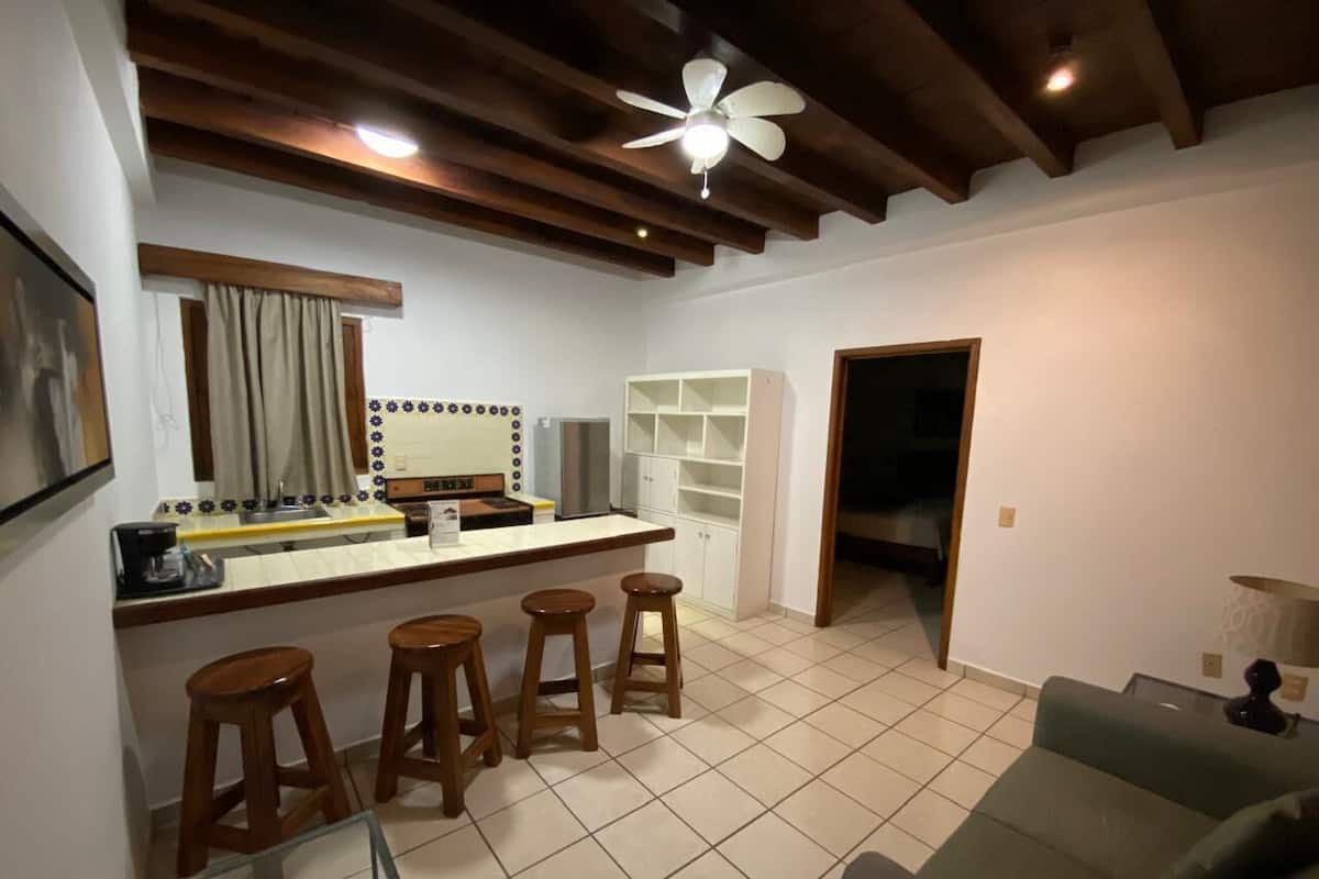 Une cuisine avec un coin bar, des tabourets, un ventilateur de plafond et une porte-fenêtre donnant sur une autre pièce.