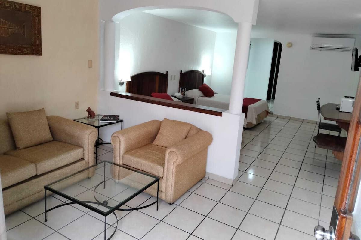Un salon avec un canapé, un fauteuil et une table basse. Au fond de la pièce, on aperçoit un lit avec un miroir au-dessus.