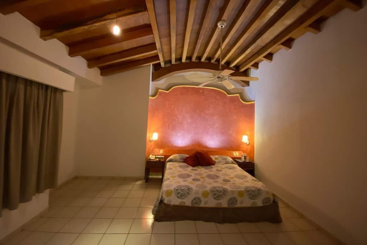 Une chambre à coucher comprenant un lit, deux tables de chevet, un ventilateur de plafond et une fenêtre avec des rideaux.