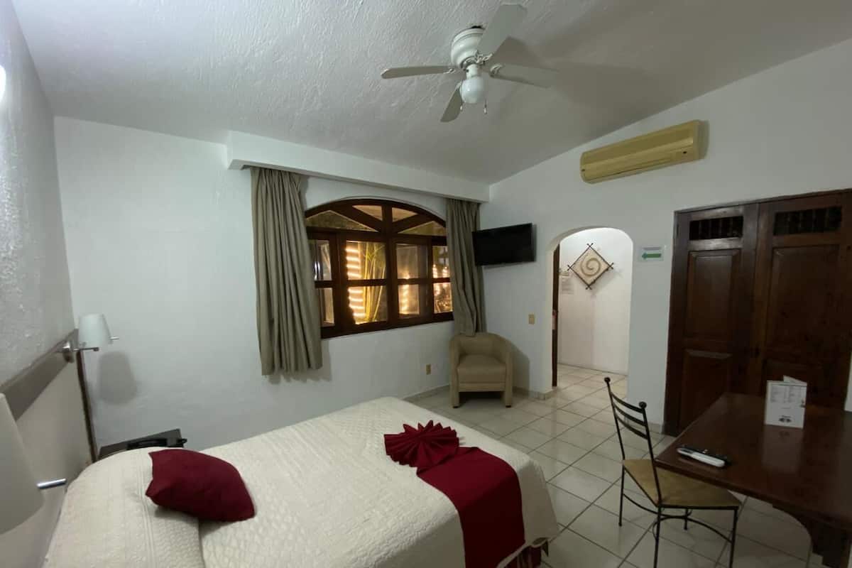 Une chambre d’hôtel avec un lit, une chaise, une table, un ventilateur de plafond et une fenêtre avec des rideaux.