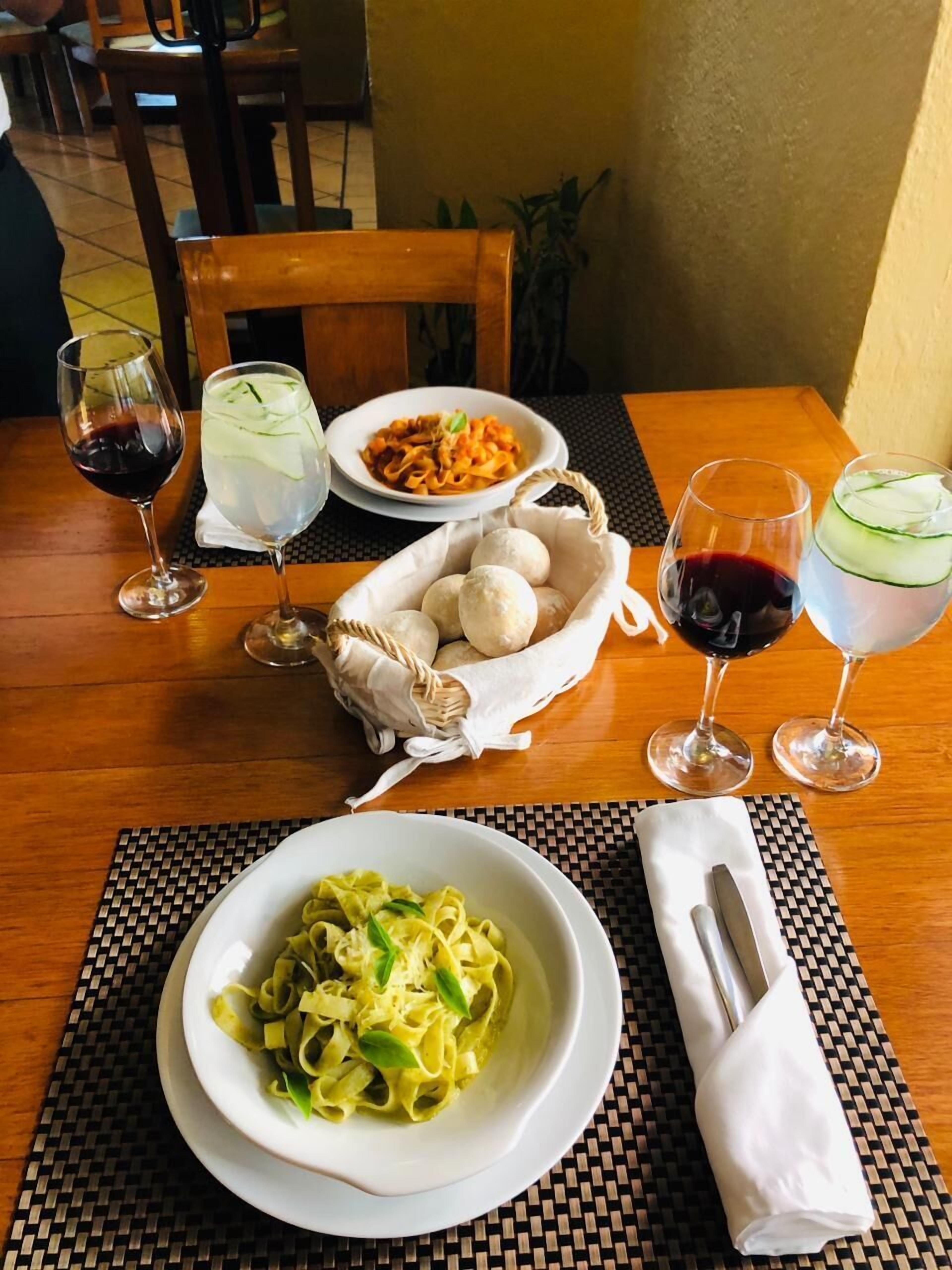 A table with a plate of pasta, a basket of bread rolls, and glasses of red wine and iced drinks.