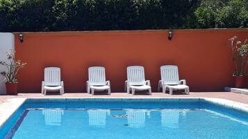 Una piscina con cuatro sillas reclinables blancas alineadas frente a un muro rojo, rodeada de follaje.
