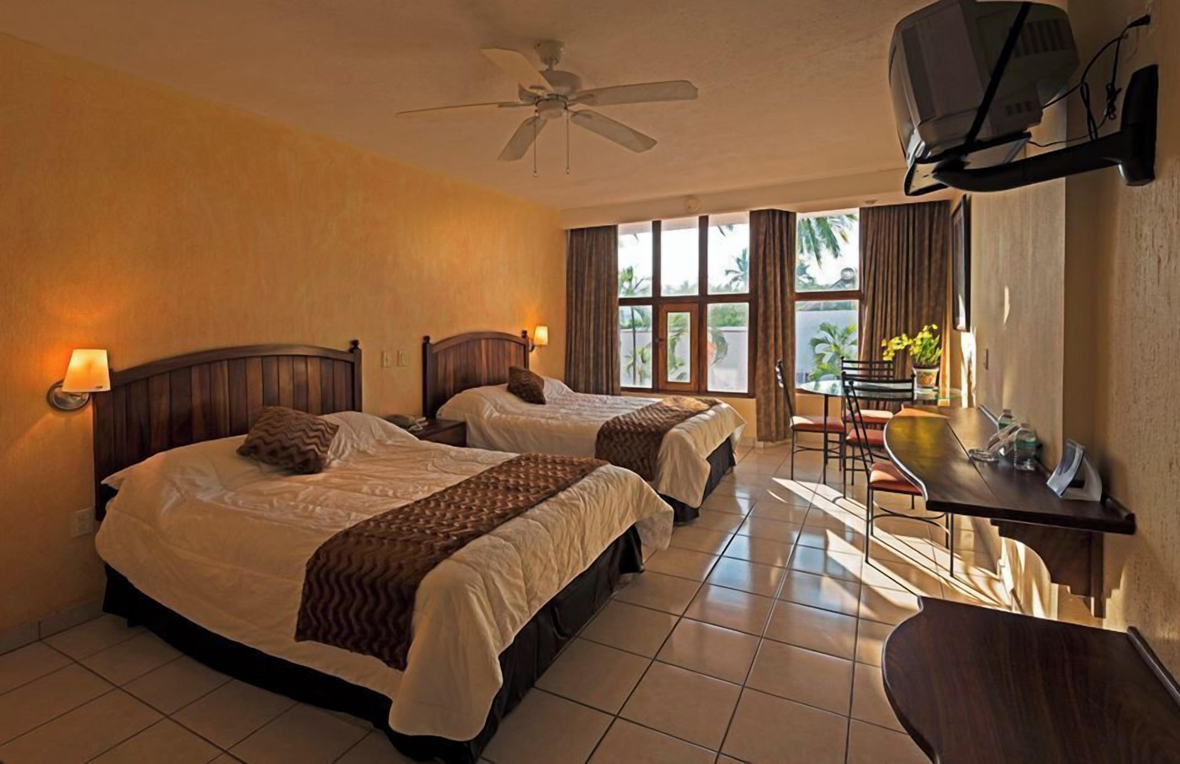 A hotel room with two beds, a desk, a chair, a television, and a ceiling fan.
