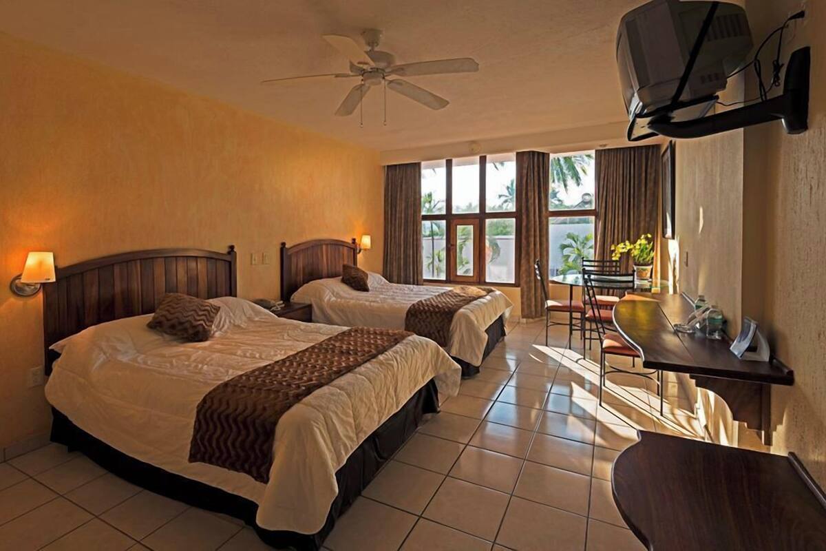 Une chambre d’hôtel avec deux lits, un bureau, une chaise, une télévision et un ventilateur de plafond.