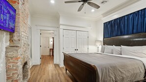 A bedroom with a wooden floor, a bed with a headboard, a ceiling fan, and a TV mounted on a brick wall. - Historic Shotgun, Near Streetcar and Superdome (New Orleans)