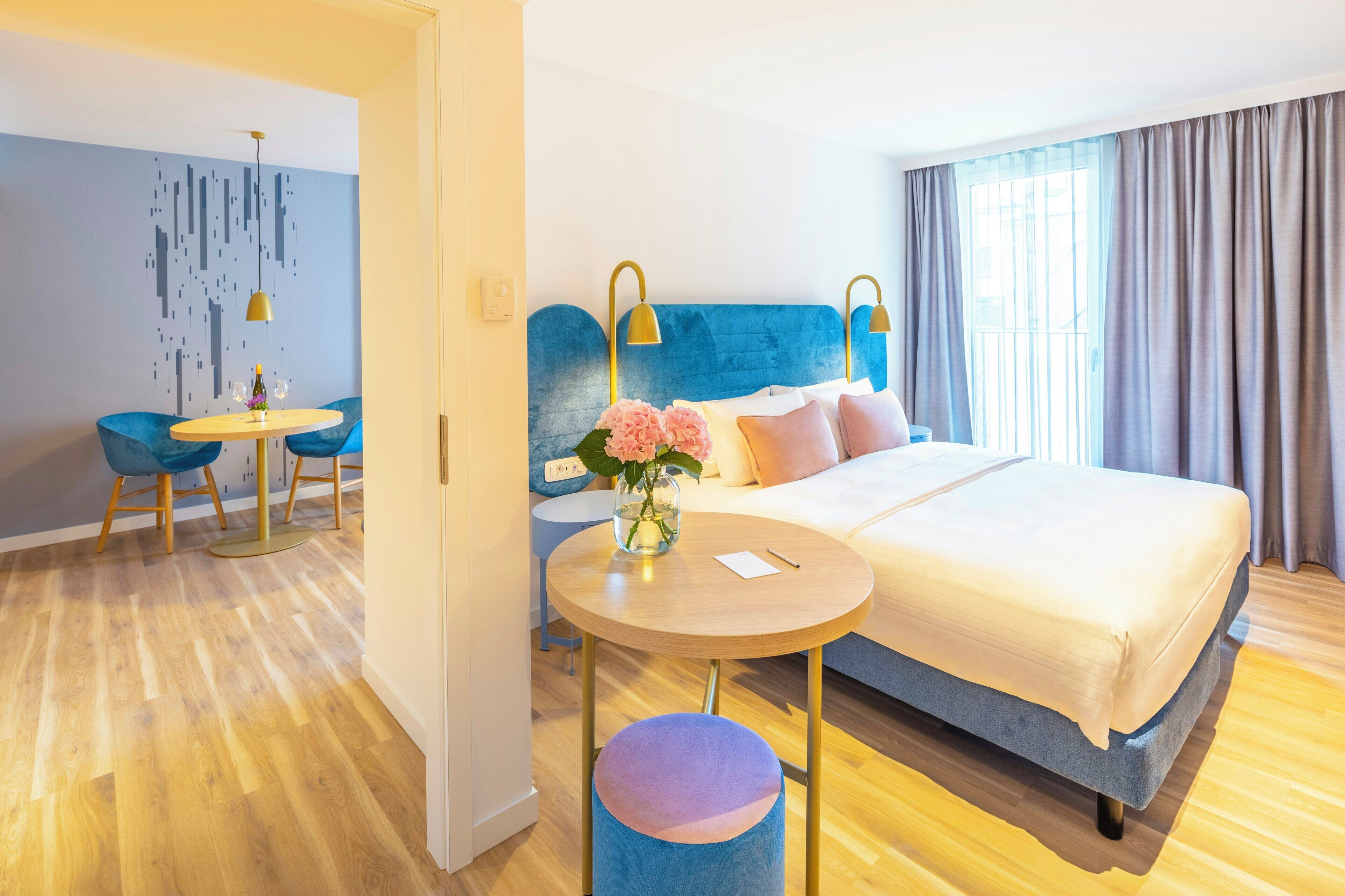 Habitación de hotel con cama, una mesa redonda, una silla azul, un jarrón con flores y una pared con diseño geométrico.