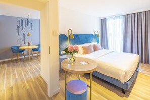 A modern hotel room with a large bed, a small round table, two chairs, a vase with flowers, and a view of the city through the window.