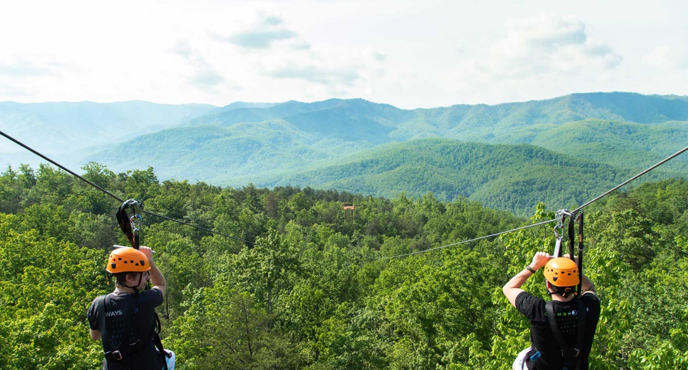 Dos personas en un tirolina con un paisaje montañoso cubierto de bosque al fondo.