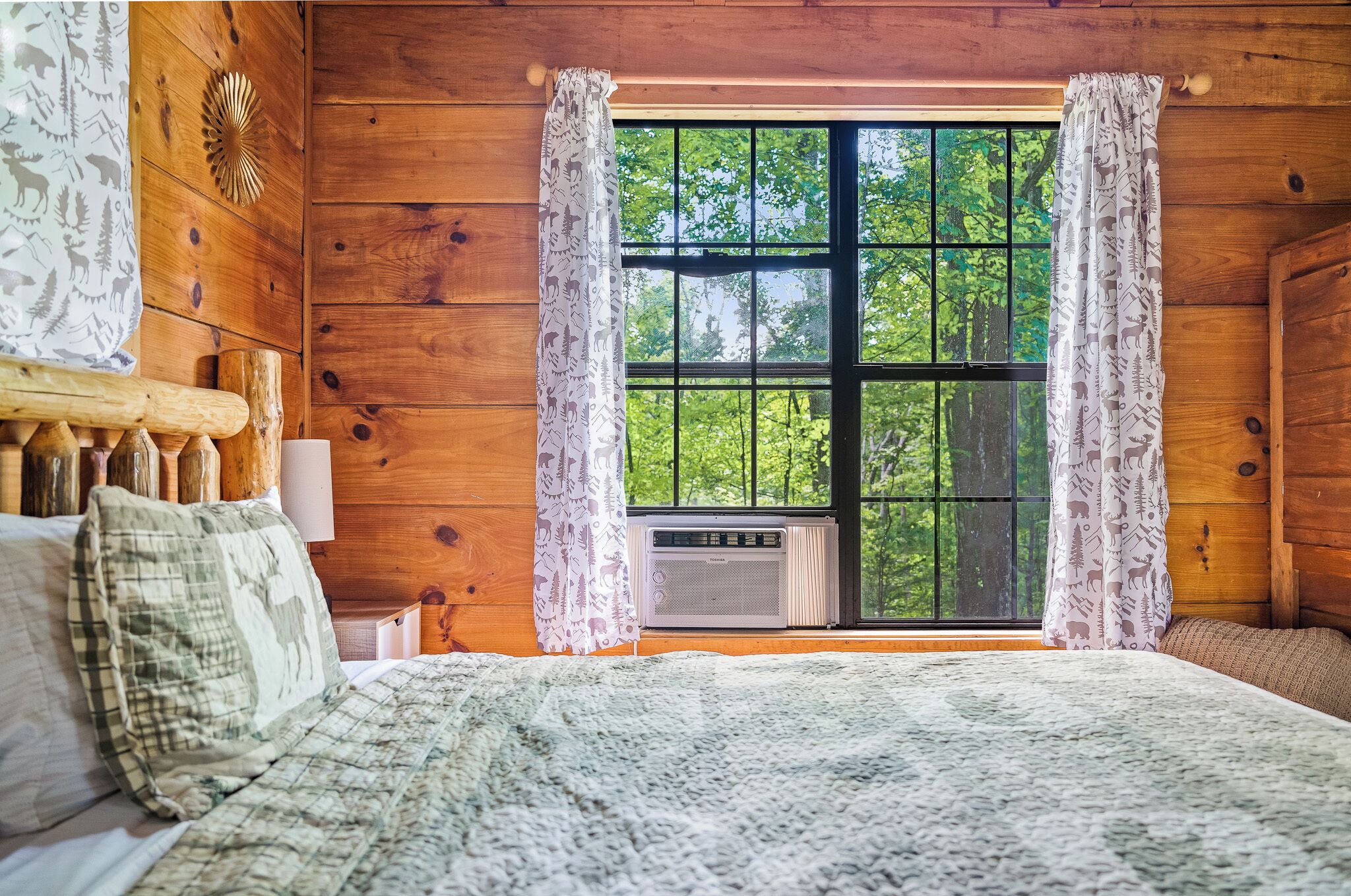 A wooden cabin bedroom with a large window, a bed with a patterned blanket, and a wall-mounted heater.