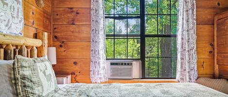 A wooden cabin bedroom with a large window, a bed with a patterned blanket, and a wall-mounted heater.
