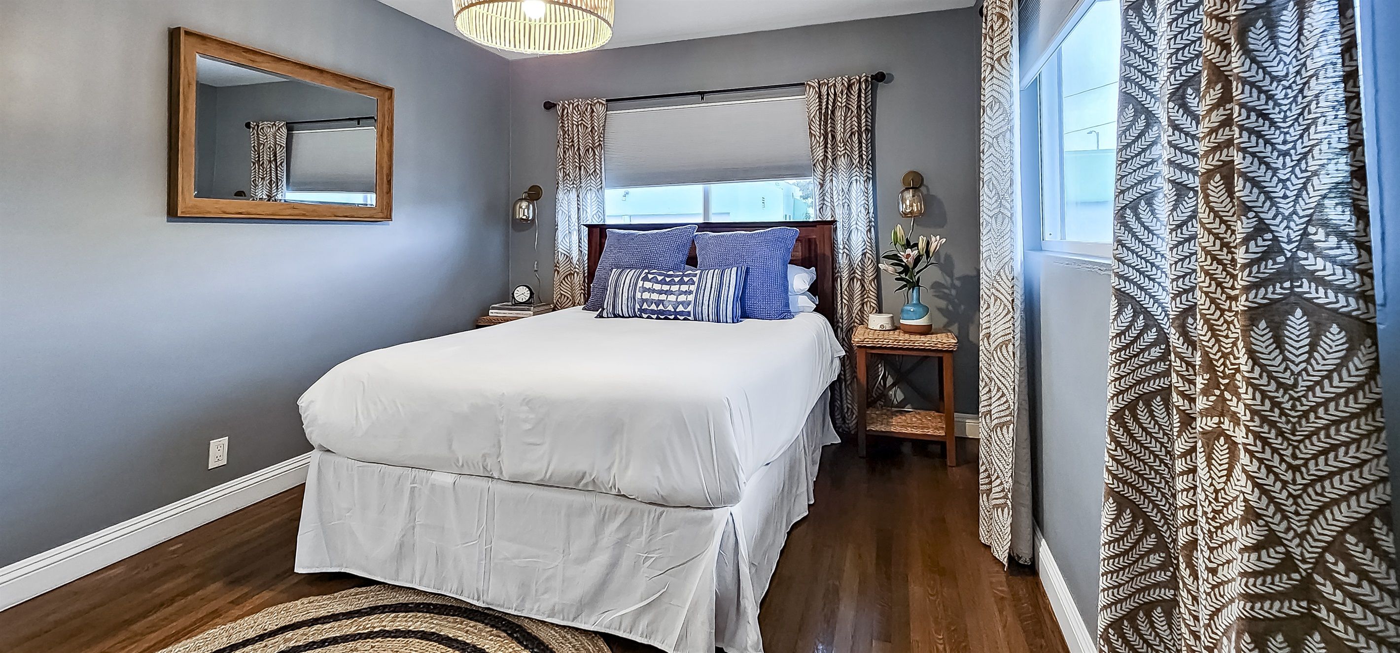 A bedroom with a bed, a nightstand, a mirror, and a window with patterned curtains.