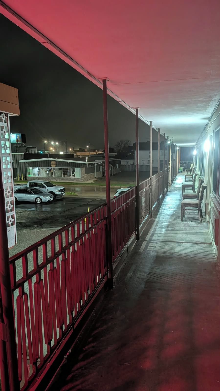 A covered outdoor area with a red railing, a paved walkway, and a row of chairs.