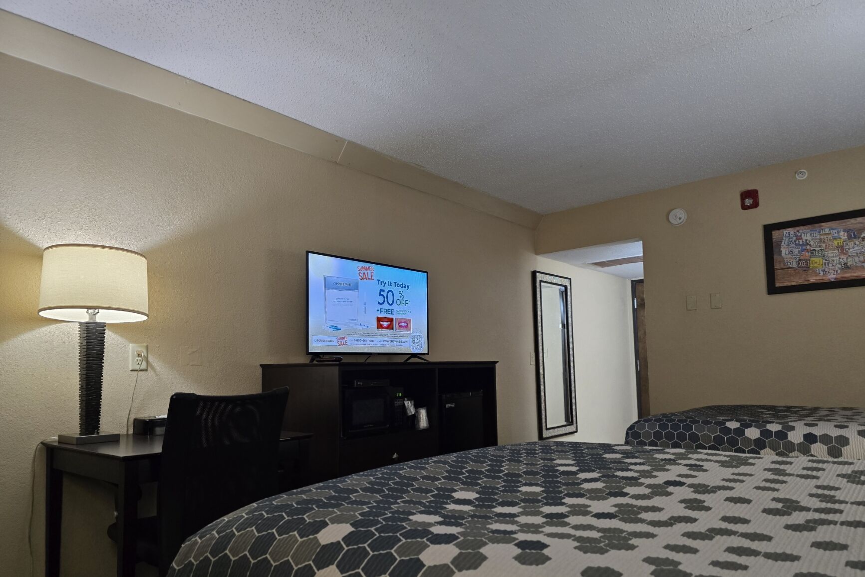 A hotel room with a bed, a TV displaying a sale, a desk with a lamp, and a mirror.
