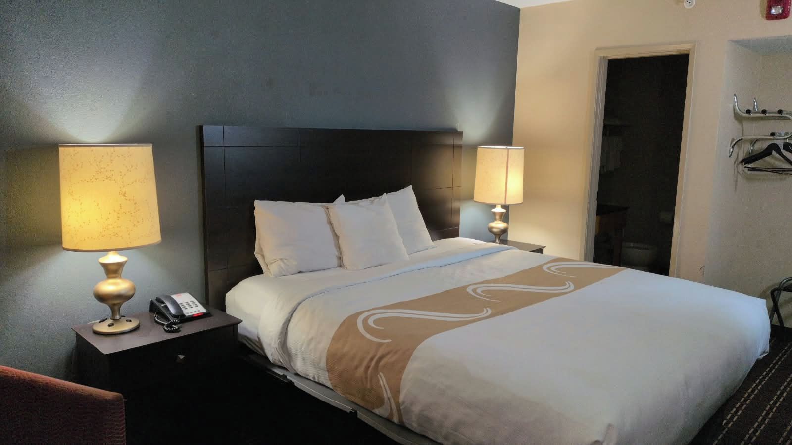 Quarto de hotel com uma cama grande, duas mesas de cabeceira com luminárias, um telefone e um porta-roupas.