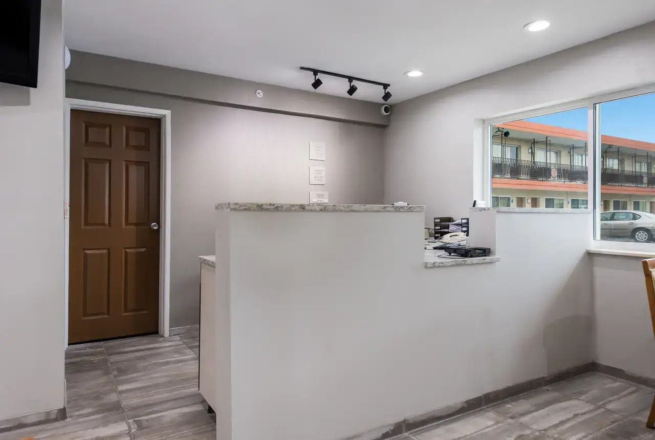 A hotel room with a wooden door, a small kitchenette, a window with a view of buildings, and a ceiling light fixture.