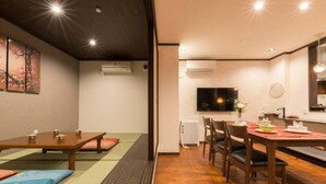 Um quarto japonês tradicional com tatames, uma mesa baixa com almofadas, uma televisão e um espaço de refeições com cadeiras e uma mesa posta para dois.