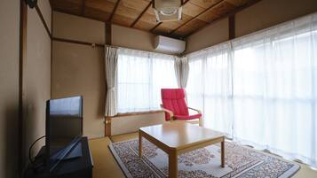 Una habitación con techo de madera, un televisor de pantalla plana sobre un mueble, una silla roja, una mesa y un ventanal con cortinas translúcidas.