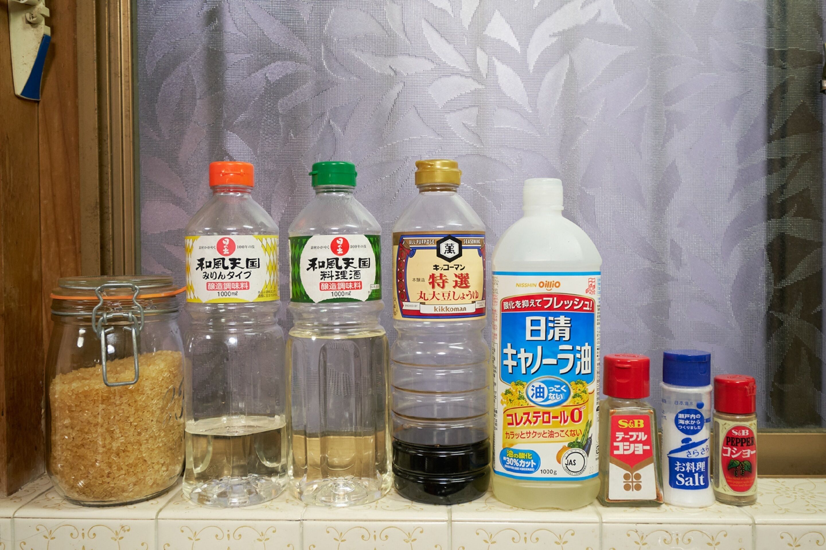A shelf with various food items including a jar of sugar, bottles of soy sauce, a bottle of rice vinegar, a bottle of salt, and two bottles of pepper.