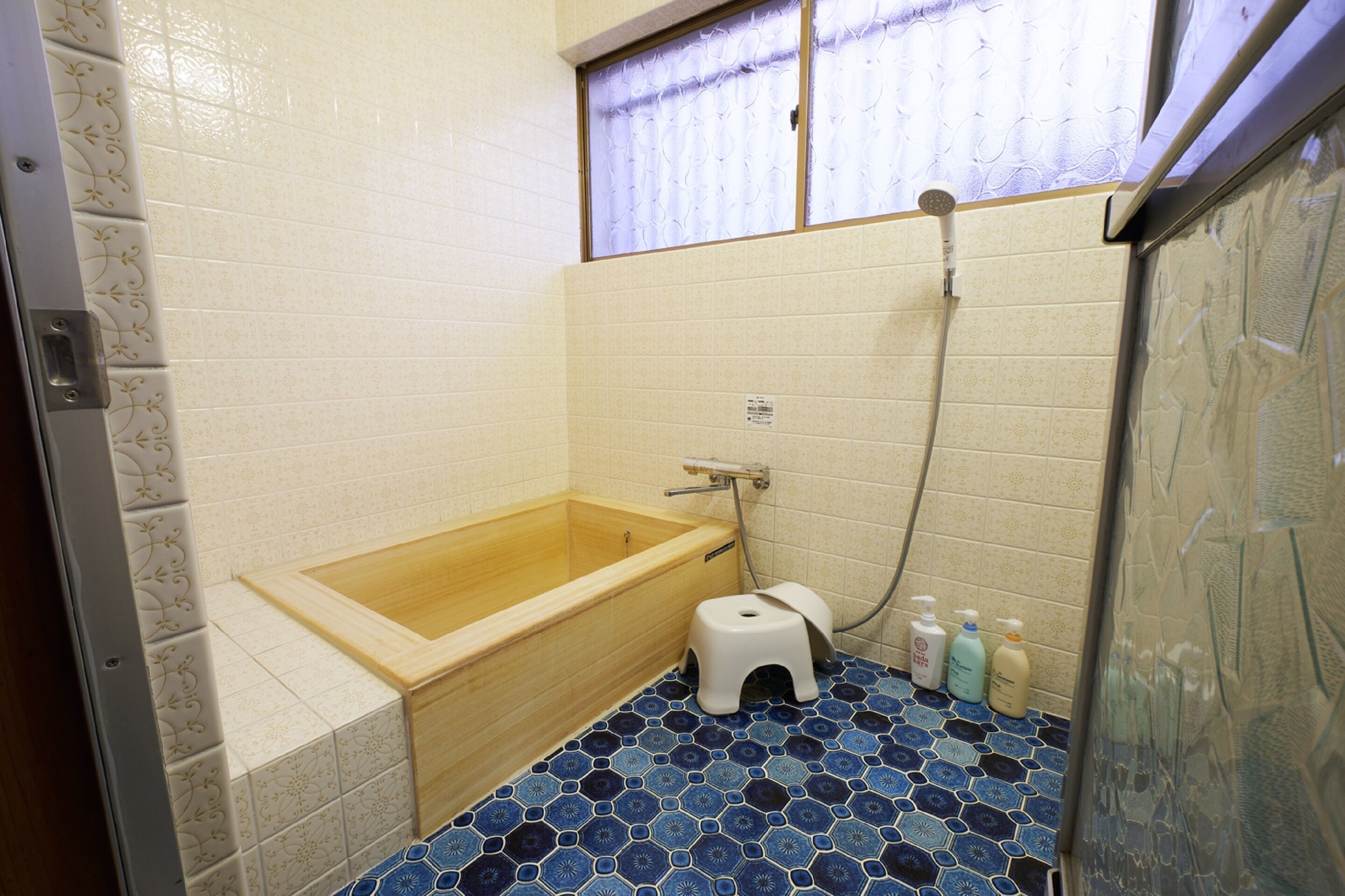 A bathroom with a wooden bathtub, a small white stool, and three bottles of toiletries on the floor.