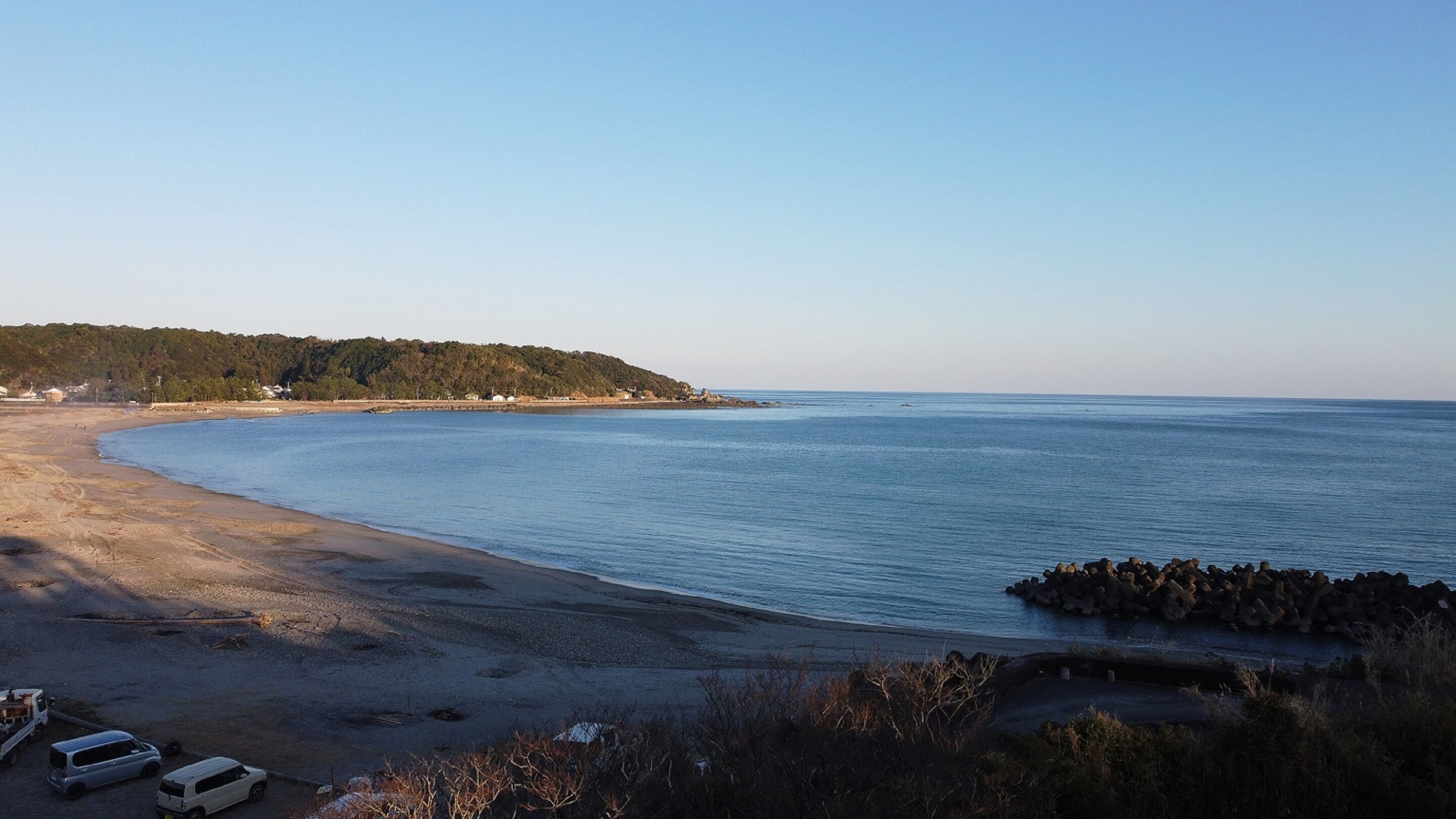A coastal beach with a sandy shore, a rocky breakwater, and a forested hill in the background.