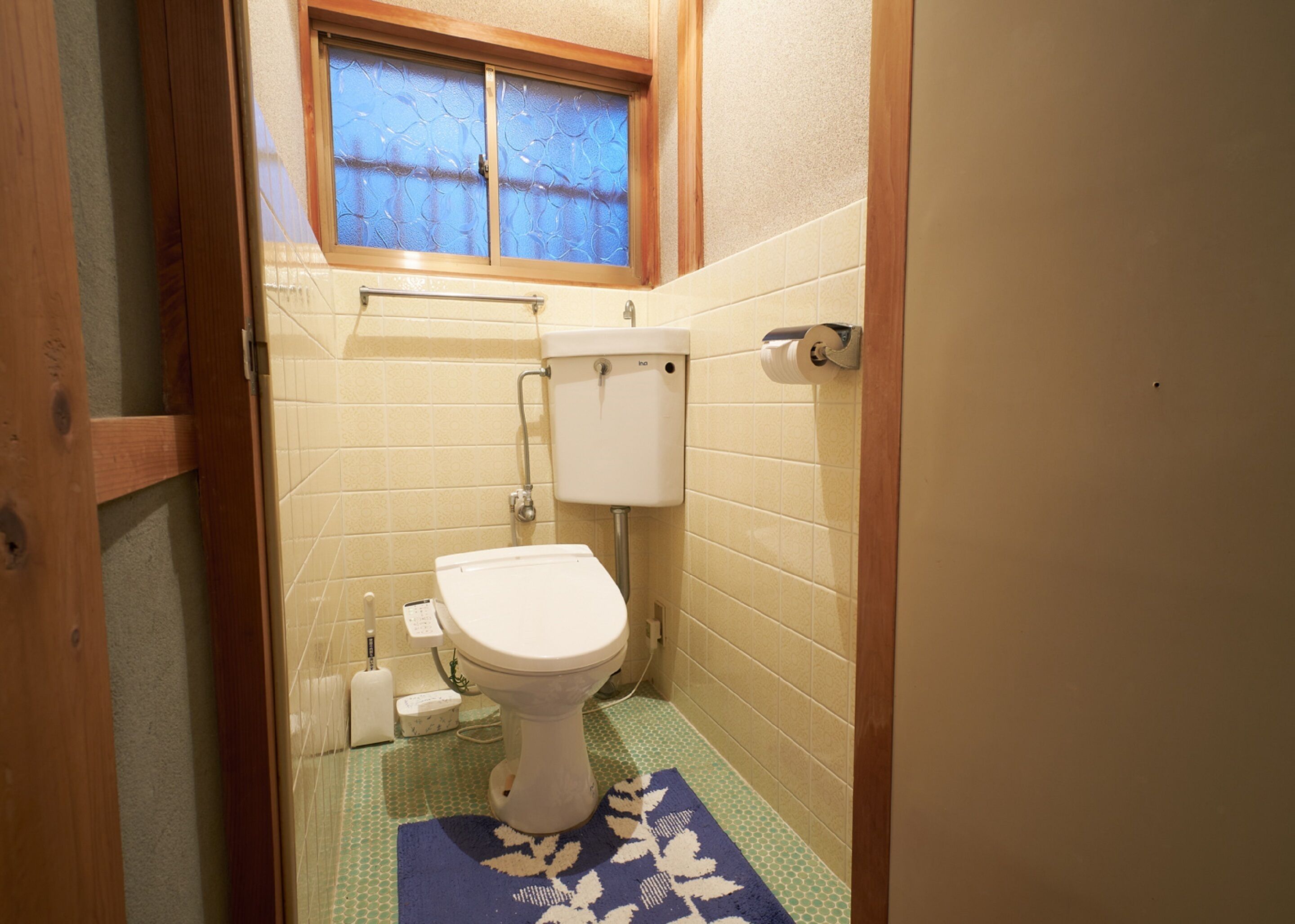 A small bathroom with a toilet, a window, and a blue patterned mat on the floor.