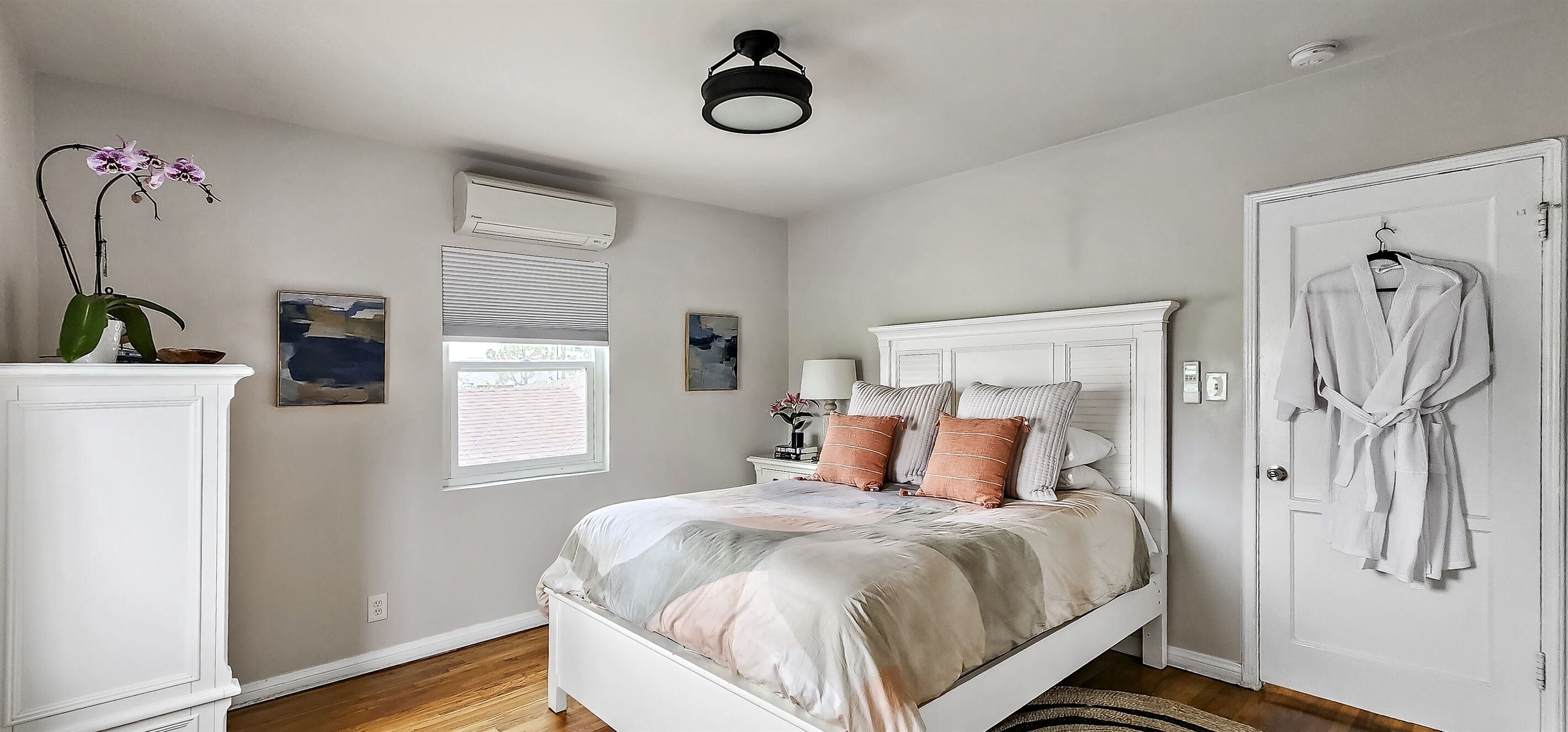 A bedroom with a bed, a white wardrobe, a door with a robe hanging, a window with blinds, and an air conditioning unit.