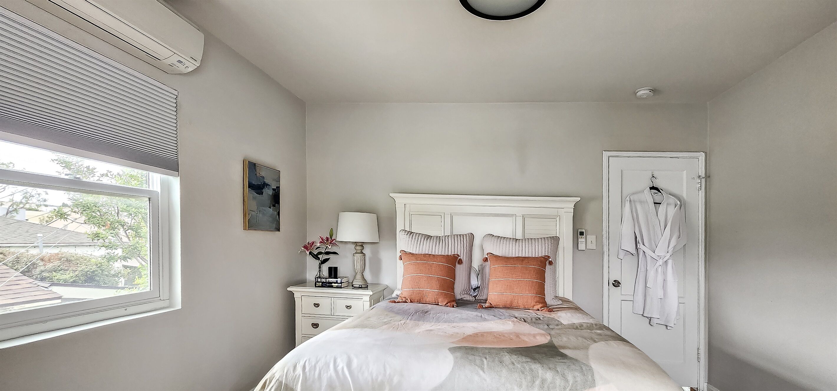 A bedroom with a bed, a nightstand with a lamp, a framed picture, and a door with a robe hanging.