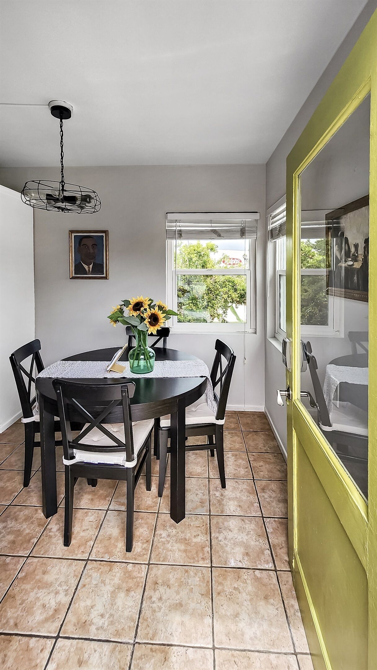 Un coin repas avec une table ronde, quatre chaises, un vase avec des tournesols, un ventilateur de plafond, un tableau encadré et une fenêtre avec des stores.