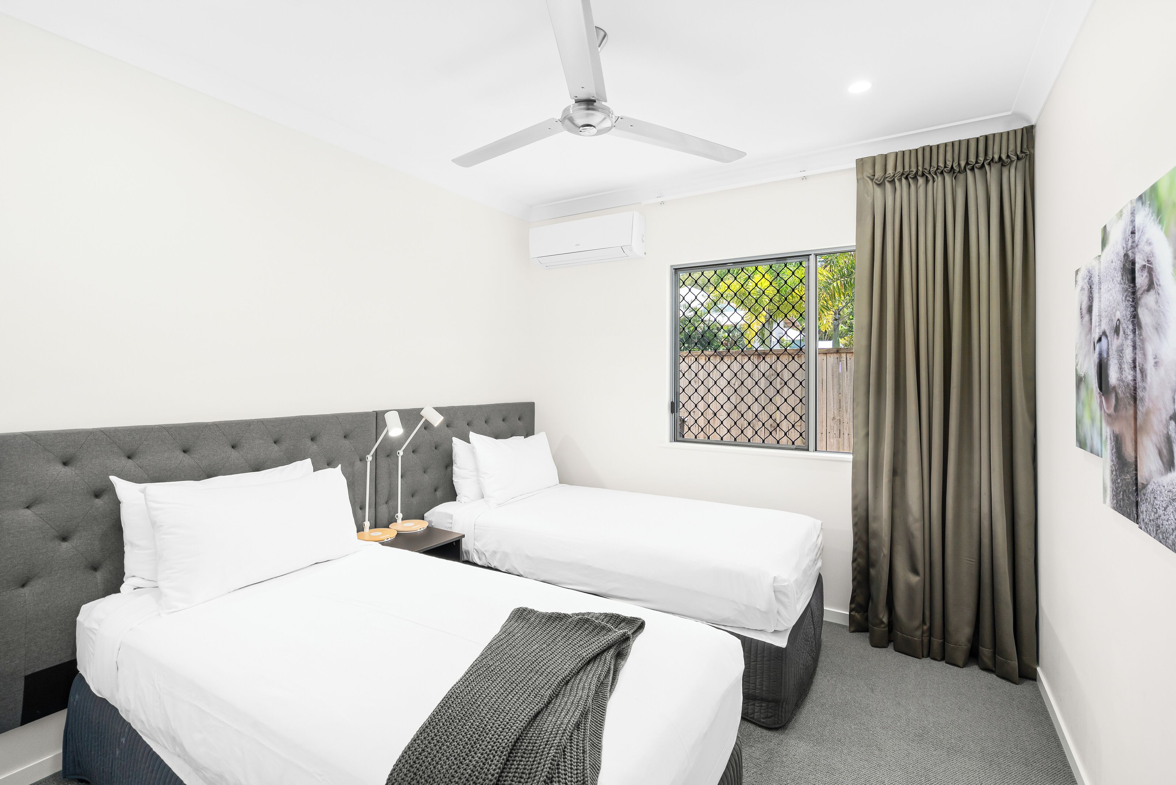 Une chambre d’hôtel avec deux lits, un ventilateur de plafond, un appareil de climatisation, une fenêtre avec des rideaux et un tableau accroché au mur.