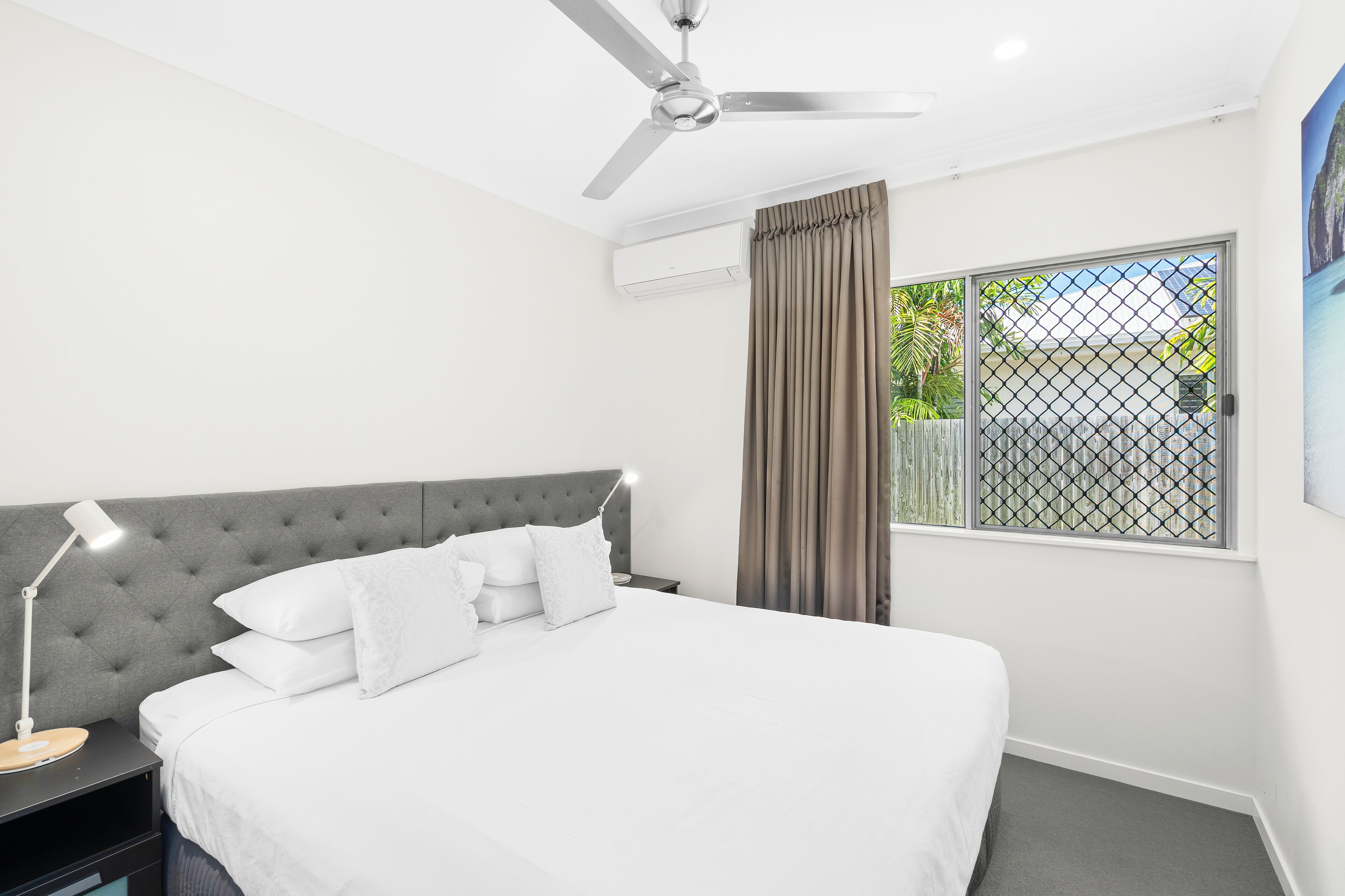 A bedroom with a bed, a nightstand, a desk lamp, a ceiling fan, an air conditioning unit, a window with curtains, and a view of greenery outside.
