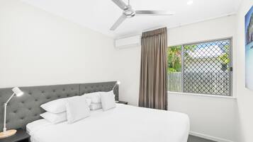 A bedroom with a bed, a nightstand, a desk lamp, a ceiling fan, an air conditioning unit, a window with curtains, and a view of greenery outside.